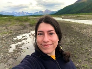 woman with dark hiar pulled back, smiling at camera, backdrop of braided river bed and mountains behind her